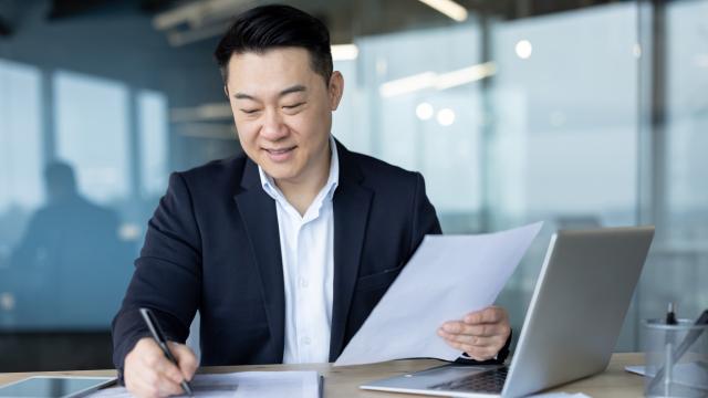 Individual looking through paperwork at table with laptop open.