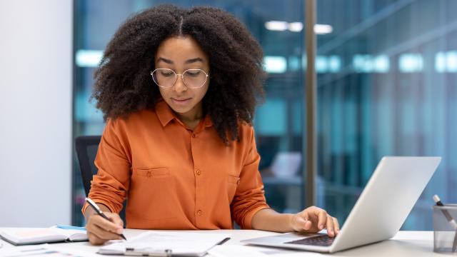 Individual sitting at table looking over paperwork and has a laptop open.
