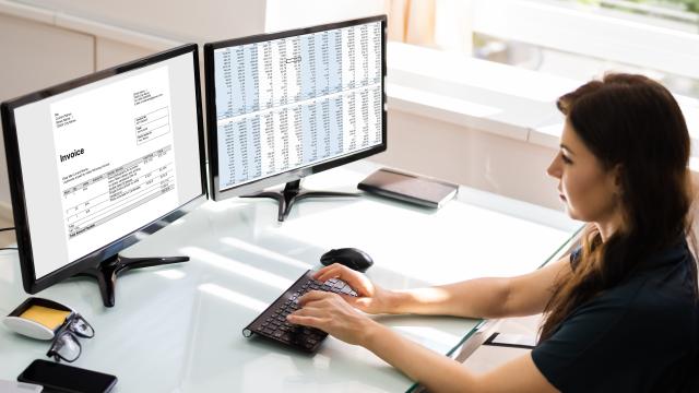 Person sitting at desk with dual monitors and reviewing an invoice and financial data