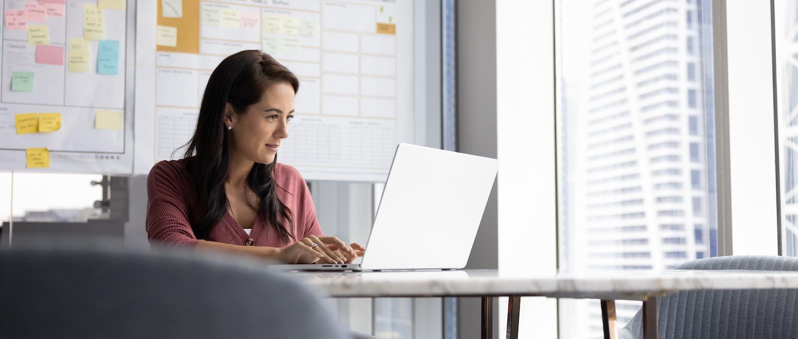 Individual sitting at table looking at laptop with post it notes and work boards behind them